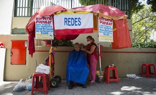 Ana Urdaneta, une coiffeuse de 64 ans, désigne fièrement une affiche du défunt président Hugo Chavez qui orne son "salon de coiffure populaire" aux tarifs "solidaires" improvisé sous des parasols rouges en pleine rue à Caracas.