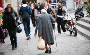 Une personne âgée dans une rue piétonne, avec un cabas et une béquille. Illustration sur la vieillesse.