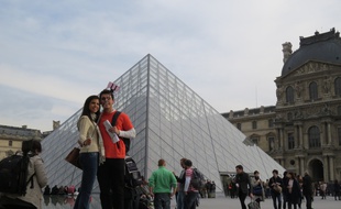 Deux touristes se prenant en portrait devant la pyramide du Louvre aidés d'une perche à selfie.