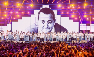 Artistes et bénévoles des Enfoirés sur la scène de l'Arkéa Arena devant le portrait de Coluche.