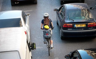 Une femme sur un vélo, avec un porte bébé, dans la circulation. Casque.