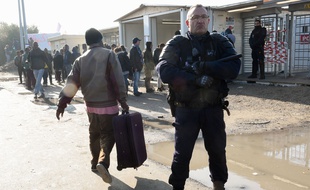 Calais (Pas-de-Calais), le 23 octobre 2016. Un migrant chargé d'une valise passe derrière un policier chargé de la sécurité dans la «jungle».