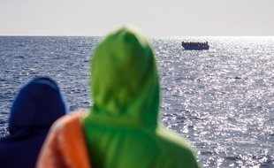 Deux migrants secoururs par un navire regardent une embarcation à secourir en Méditerranée.